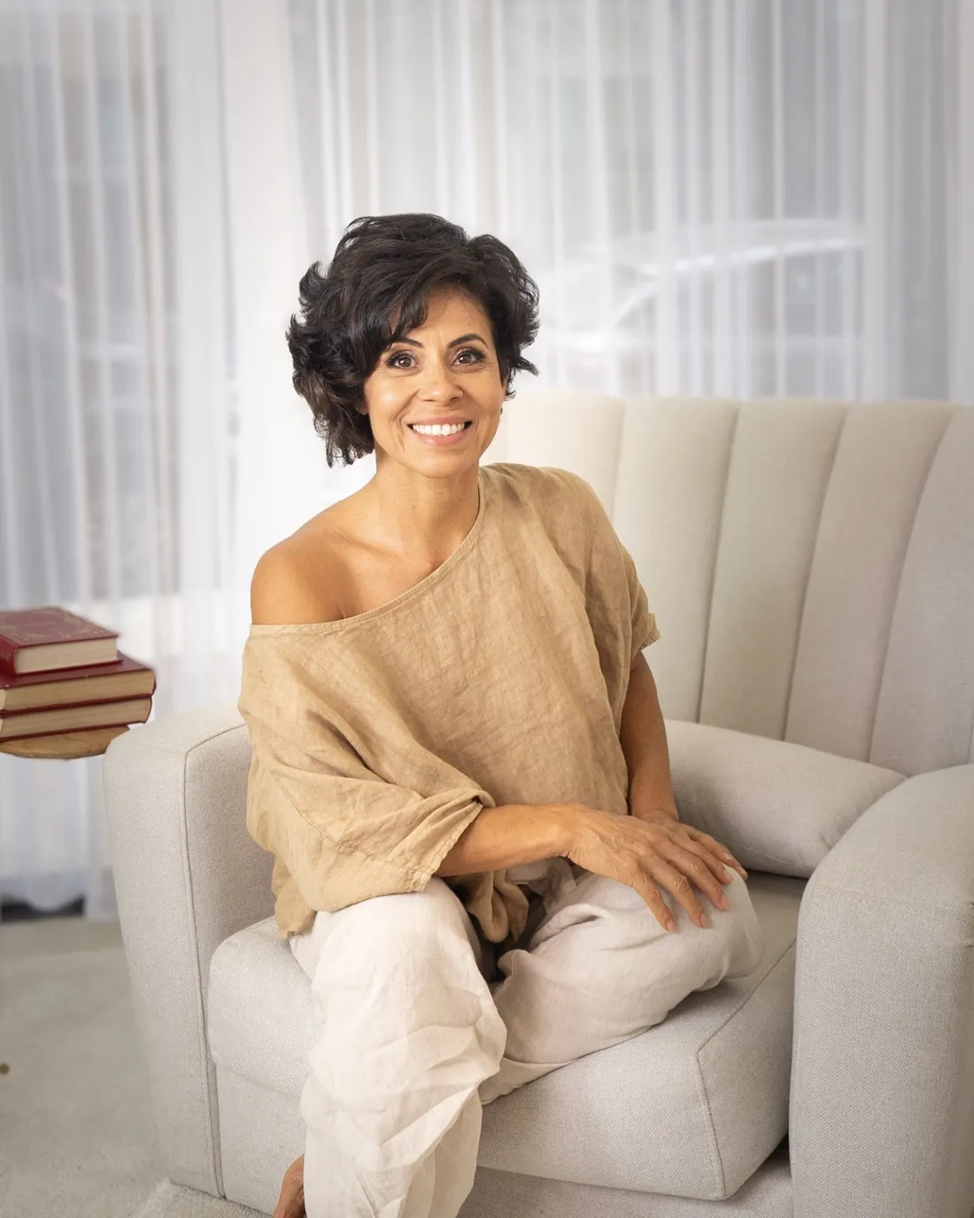 Marlene Faustino, com blusa de linho bege, sentada em uma poltrona clara, sorrindo