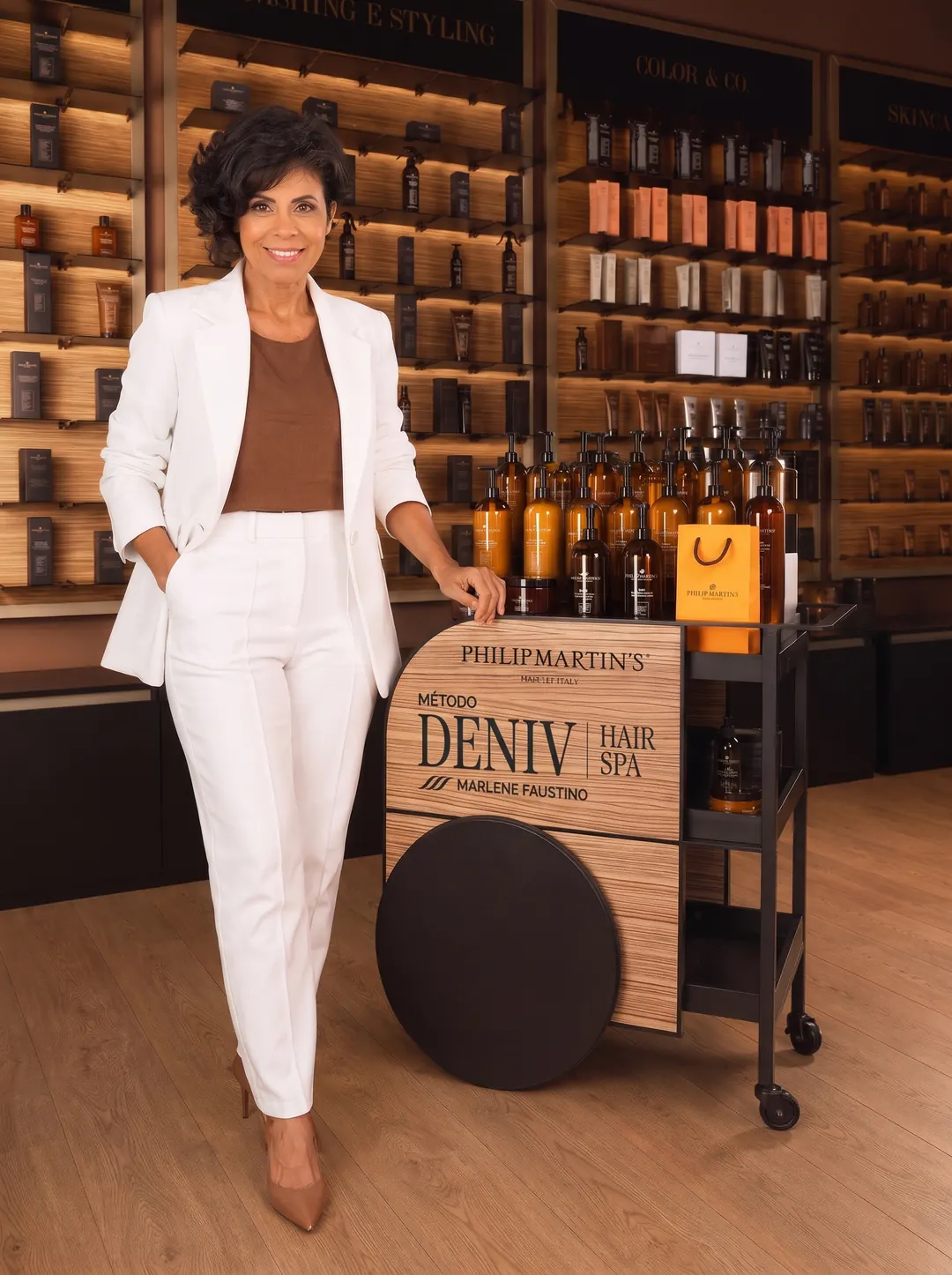 Marlene Faustino de terno branco, na loja da Philip Martin’s, ao lado do carrinho do Método DENIV Hair Spa com os produtos da marca